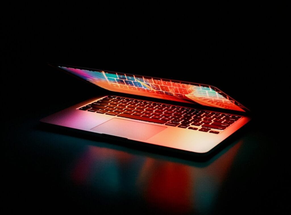 Laptop partially opened, emitting vibrant multicolored lights in dark setting.